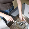 A hand is placing a flexible silicone sink splash guard with a suction base into a stainless steel sink.