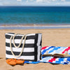 A black and white striped canvas shoulder bag resting on the sand with an open book and a blue towel.
