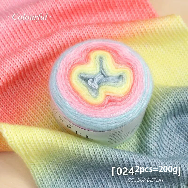A roll of soft, colorful gradient cake yarn in pink, yellow, and blue tones displayed on a yellow fabric.