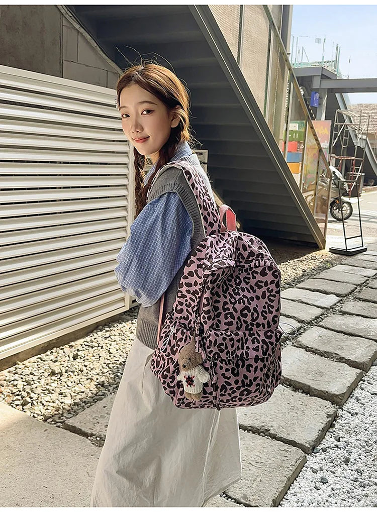 A young woman wearing a pink leopard print nylon backpack and a white skirt stands outdoors near a metal structure.