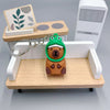 An adorable capybara keychain charm with a green hood and brown body is displayed on a wooden tray.