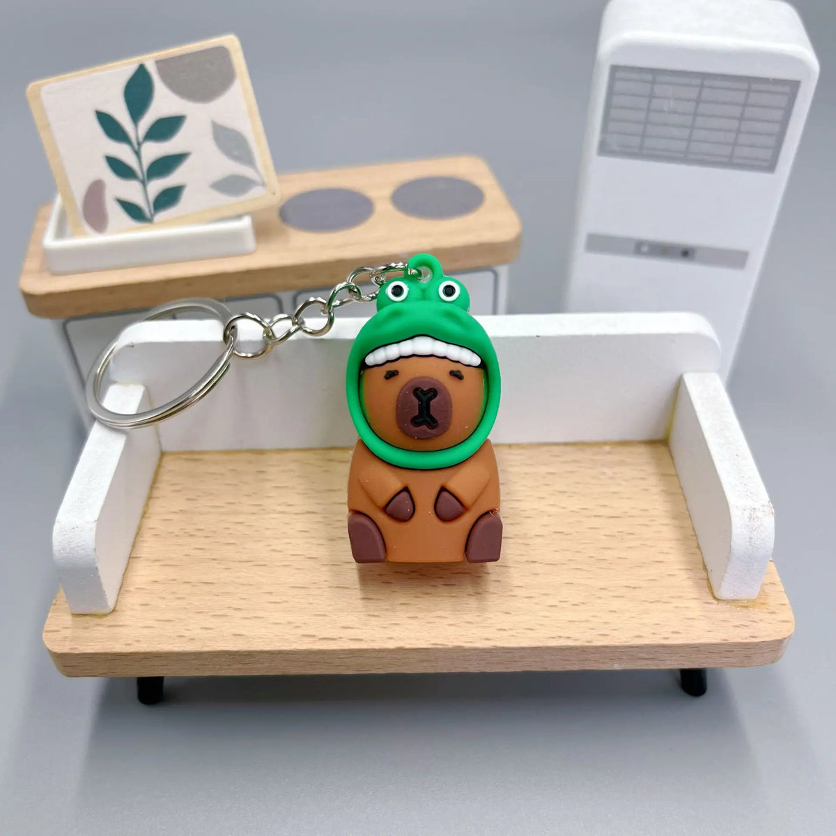 An adorable capybara keychain charm with a green hood and brown body is displayed on a wooden tray.