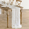 A rustic white cotton gauze table runner draped over a wooden dining table set for an event.