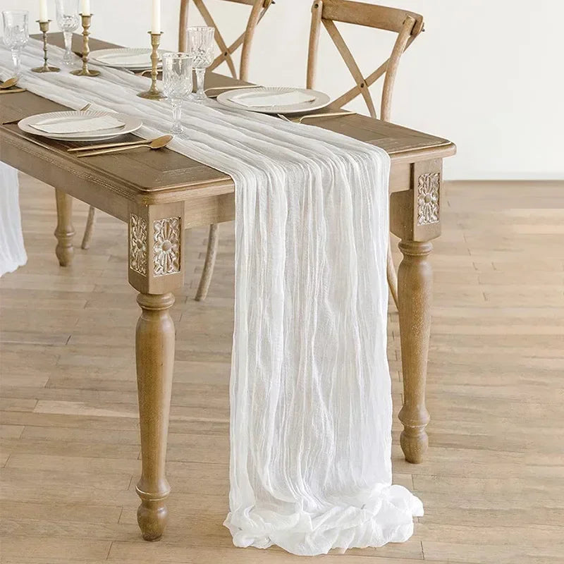 A rustic white cotton gauze table runner draped over a wooden dining table set for an event.