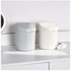 A white compact covered desktop trash bin with a flip lid sitting on a white shelf.