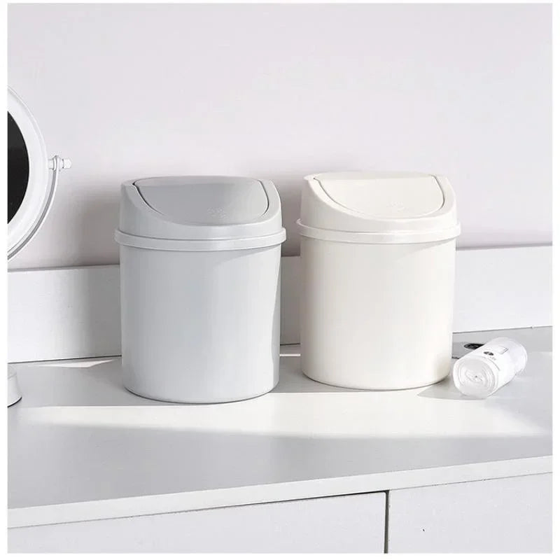 A white compact covered desktop trash bin with a flip lid sitting on a white shelf.