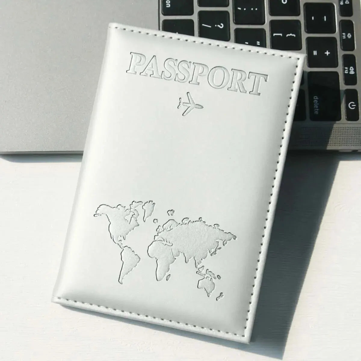 Weißes Leder-Reisepass-Halter mit silberner Weltkarte und Flugzeugsymbol auf einem grauen Laptop.
