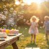 A white bubble machine is shown on a wooden table with bubbles floating in the air outdoors.