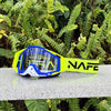 A pair of yellow and blue HD Motocross goggles with an anti-fog lens resting on a gray surface amidst green foliage.