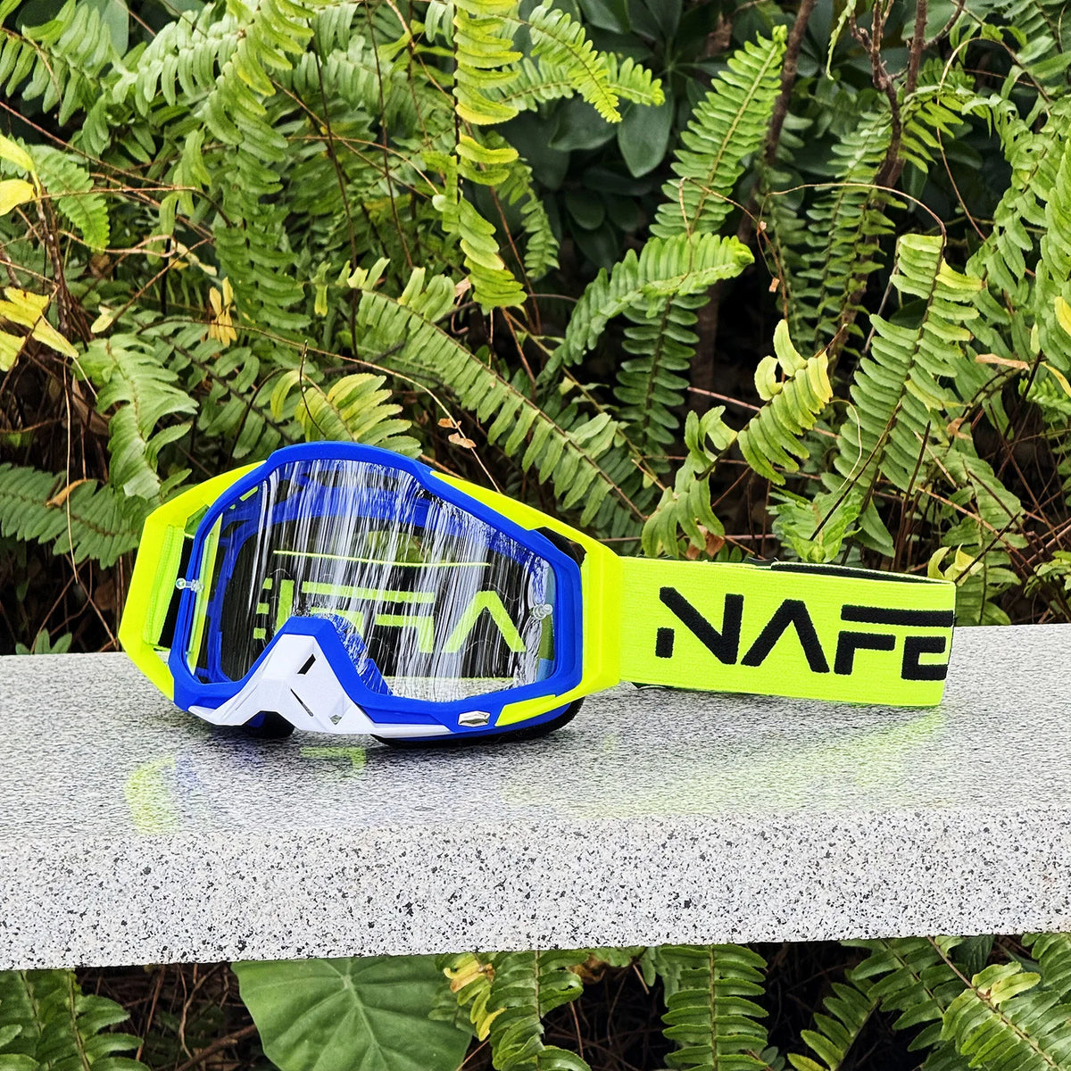 A pair of yellow and blue HD Motocross goggles with an anti-fog lens resting on a gray surface amidst green foliage.