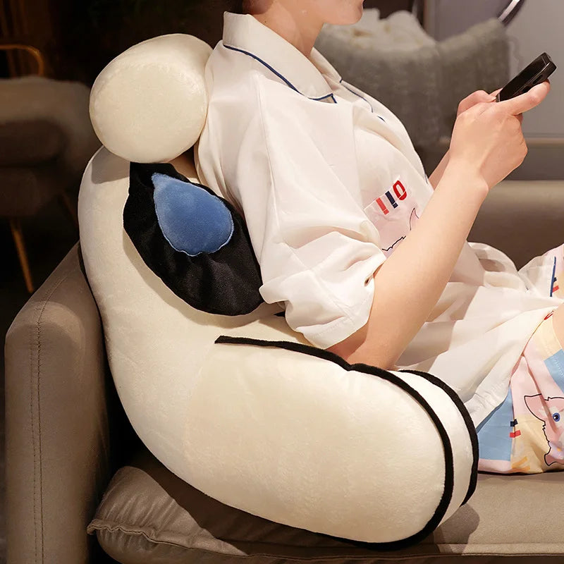 A person is relaxing on a sofa while holding a dark electronic device, with an adorable cat and dog plush backrest cushion vi