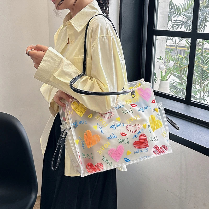 A person holds a clear transparent tote bag with a yellow and pink floral pattern and a black strap.
