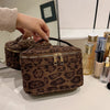 A brown leopard print cosmetic bag with a leather strap is displayed on a white surface.