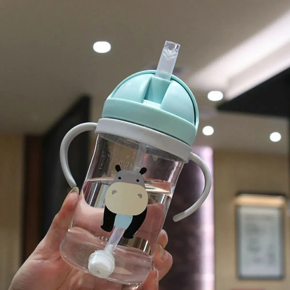 A hand holds a clear plastic sippy cup with a light blue lid and a black bear design, featuring a gravity straw.