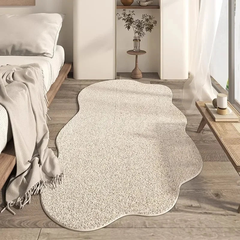 A light beige irregular area rug with a plush texture placed on a wooden floor next to a white bed and shelving unit.