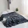A dark gray linen bed sheet spread out on a white floor with white pillows and a potted green plant in the background.