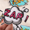 A close-up of an embroidered lettering patch featuring the word ZAP in pink and black on a white cloud shape.