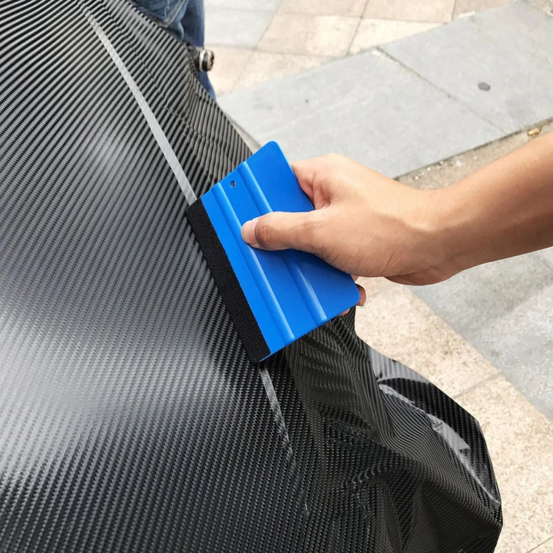 A hand uses a blue suede felt squeegee to apply a vinyl wrap or window tint onto a black surface.