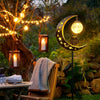 A solar moon-shaped garden light with an owl and glass ball hangs in a wooded outdoor setting.