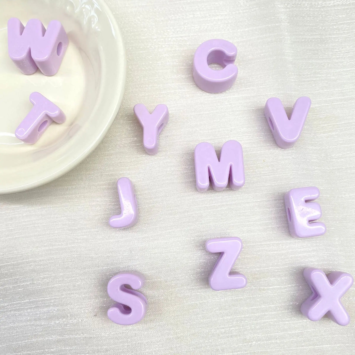 Perles en acrylique violettes formant l'alphabet éparpillées sur une surface blanche à côté d'un petit plat