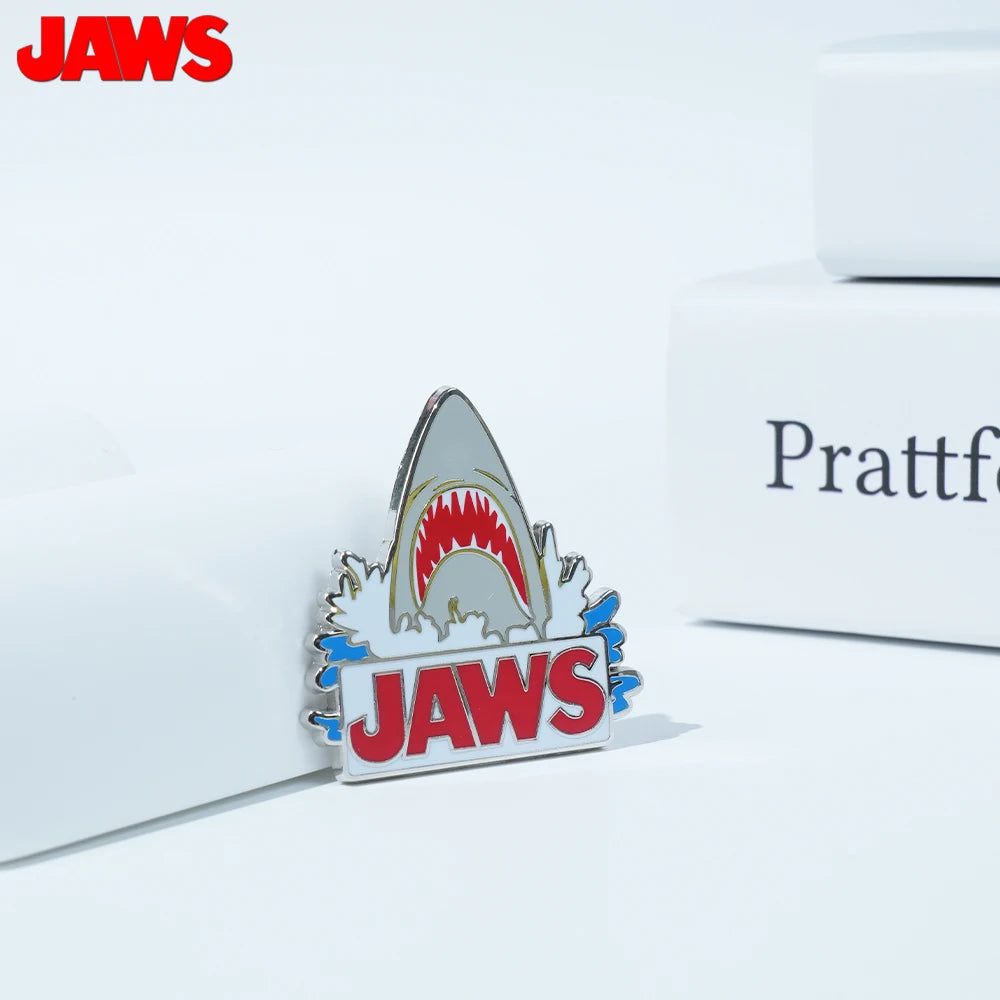 Silver enamel pin featuring a shark tooth design and the word JAWS on a white background.