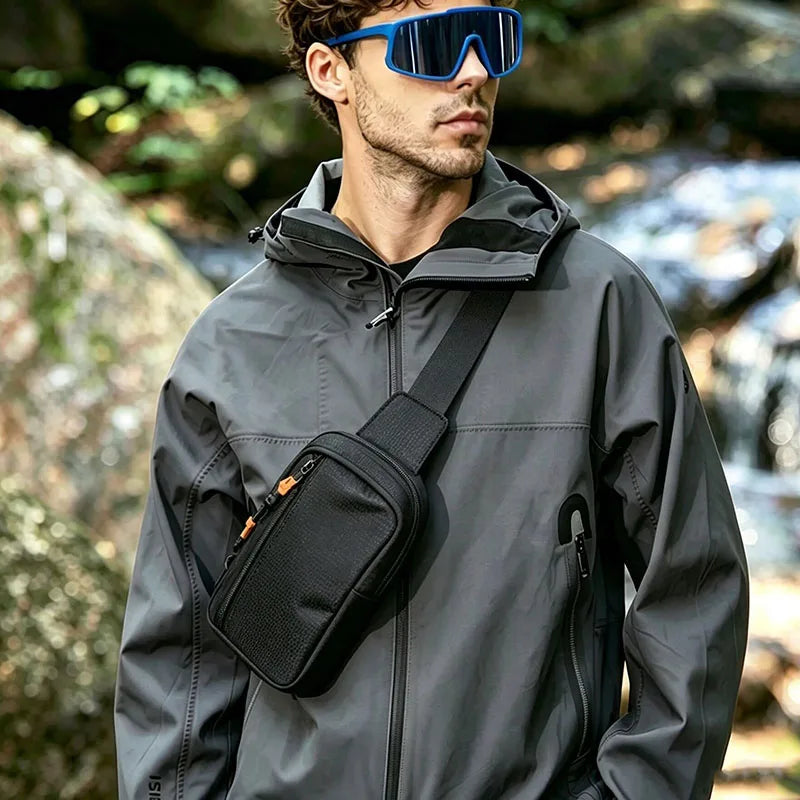 A man wearing a gray jacket and blue sunglasses holds a black sling bag in an outdoor setting.