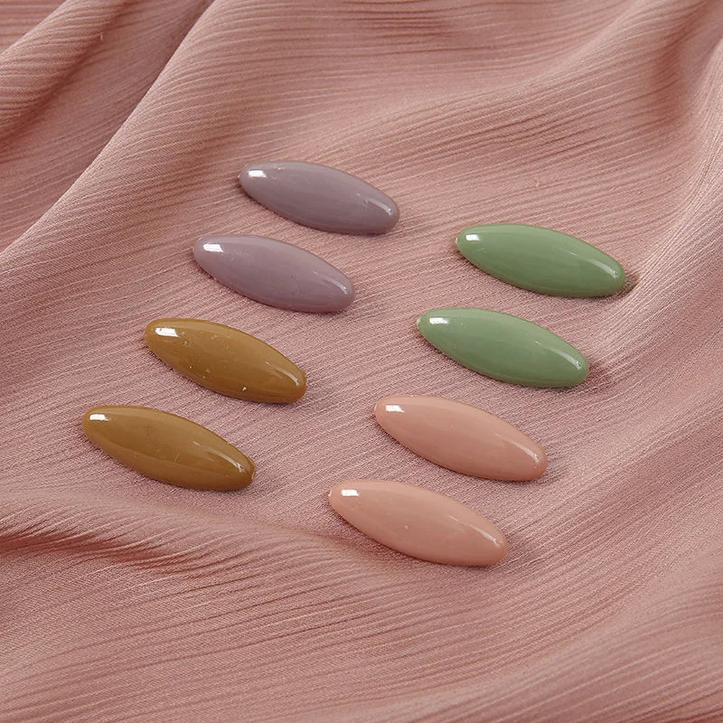 A set of eight oval solid color scarf pins in shades of lavender, mint green, yellow, and pink resting on a pink fabric.