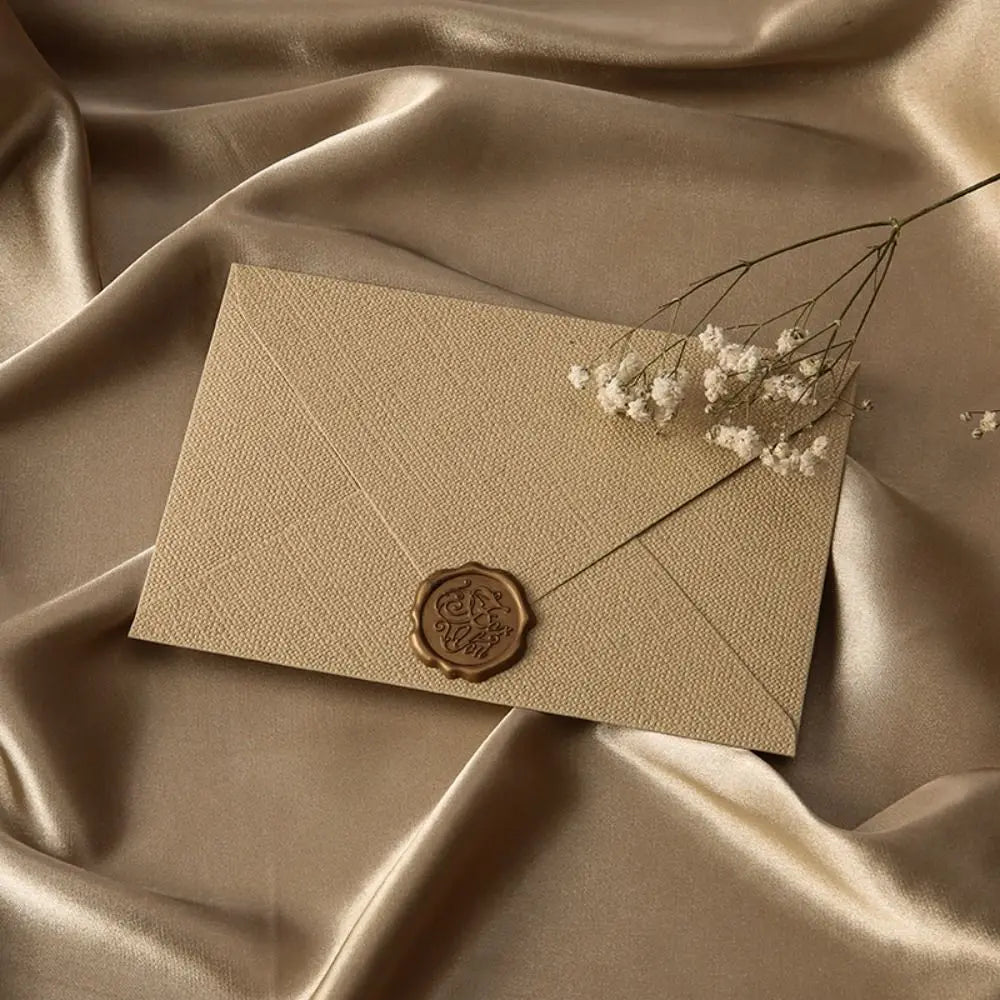 A beige linen texture envelope is displayed on draped satin fabric with a delicate sprig of baby's breath.