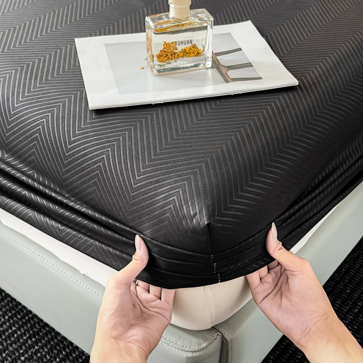 Hands are folding a black fitted sheet onto a white mattress, with an amber glass bottle and some yellow material resting on 