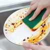 A hand is using a green and yellow double-sided cleaning sponge to wipe orange food stains off a white plate.