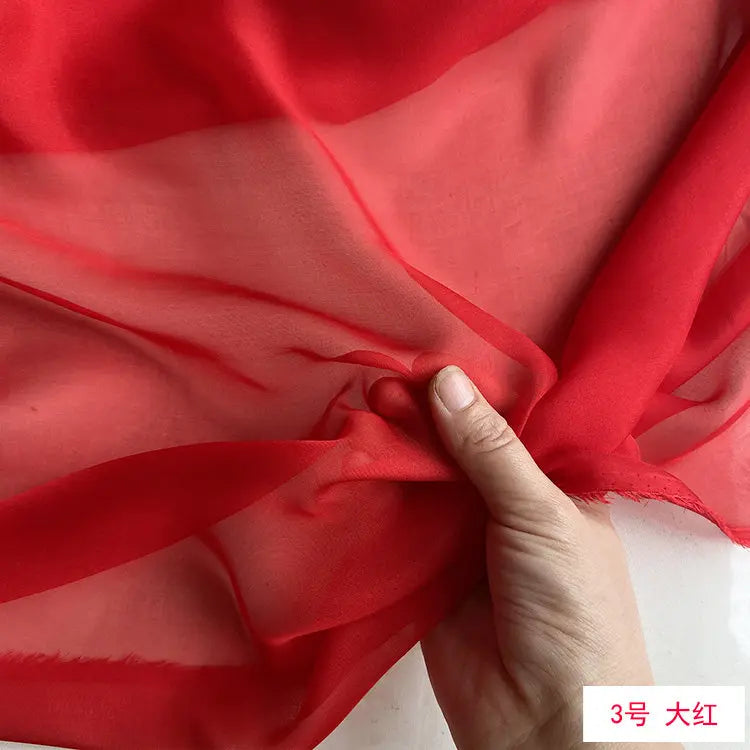 A hand is folding a piece of sheer red silk chiffon fabric, showcasing its lightweight and translucent quality.