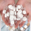 A hand holds several small, white, compressed cleansing towels packaged in clear plastic bags.
