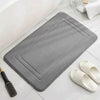 A gray plush memory foam bathroom mat with a rectangular design is shown next to a pair of white slip-on shoes.