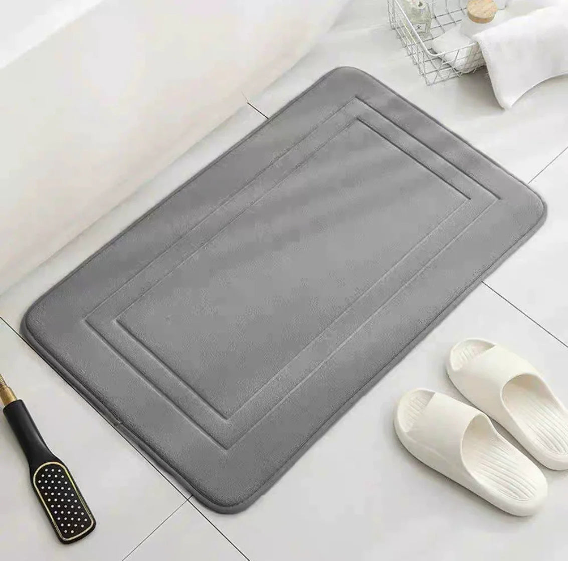 A gray plush memory foam bathroom mat with a rectangular design is shown next to a pair of white slip-on shoes.