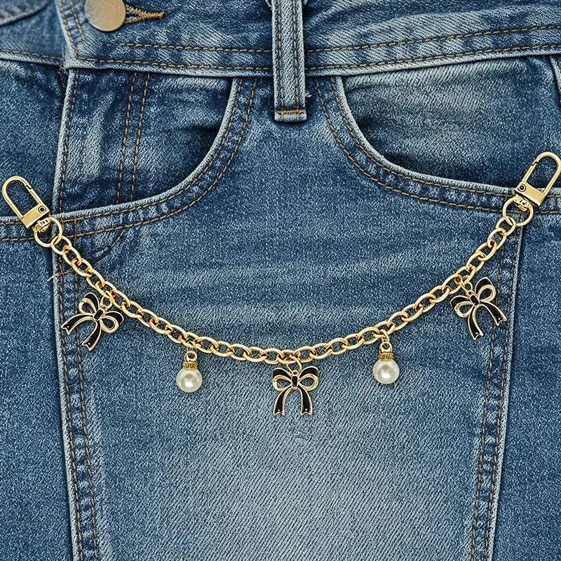 A close-up of a gold alloy chain with pearl and bow decorations attached to a denim jean pocket.