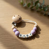 A wooden pacifier clip featuring purple beads and the name BABY carved into small wooden pieces.