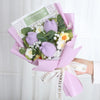 Ein Strauß handgefertigter Häkelblumen in lila und grün, eingebettet in rosa Papier