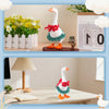 A white goose figurine wearing a red floral collar and green trim is displayed on a wooden stand.