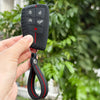 A hand holds a black silicone key fob cover with various icons, attached to a black lanyard.
