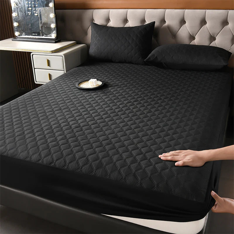 A black quilted waterproof mattress protector is shown on a bed with pillows and a bedside table.