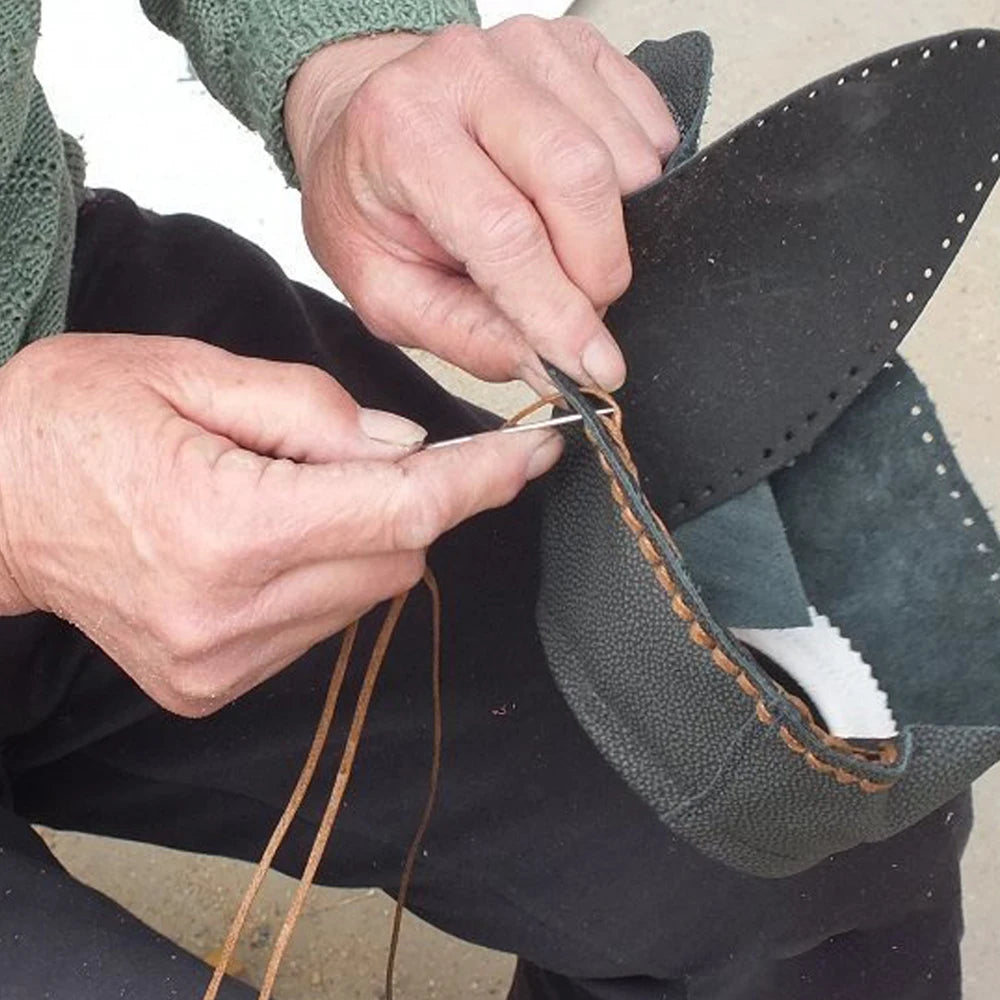 Hands are sewing a piece of dark leather fabric using a silver needle and thread.