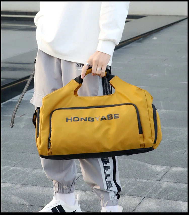 A person is holding a yellow duffel bag with a shoe pocket in an outdoor setting.