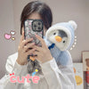 A person holds a fluffy blue penguin plush toy with an adorable hat, taking a photo with a smartphone.