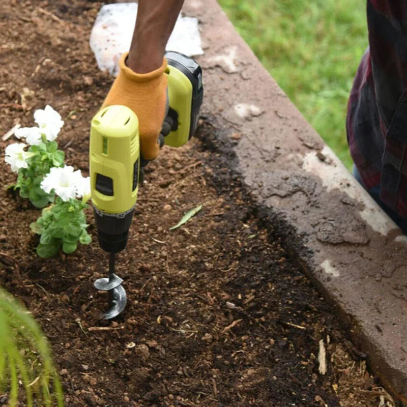 A person is using a yellow garden auger drill bit in dark soil next to a concrete edge.