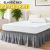 White bed with a gray ruffle skirt and white pillows on a light wood floor.