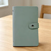 A light sage green 100-slot cash organizer binder with a snap closure resting on a wooden surface.