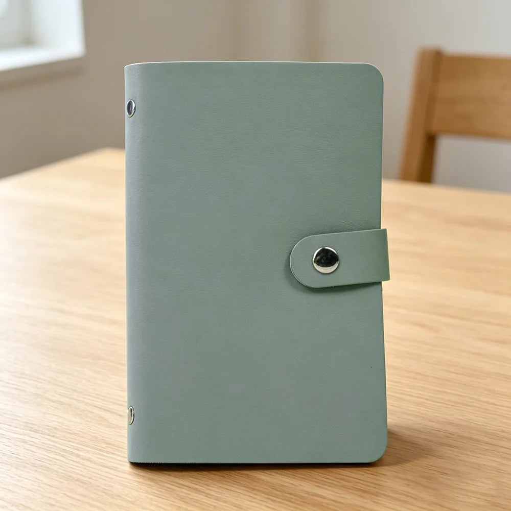 A light sage green 100-slot cash organizer binder with a snap closure resting on a wooden surface.