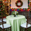 A round satin tablecloth in light green set up outdoors with wooden chairs and holiday decorations in the background