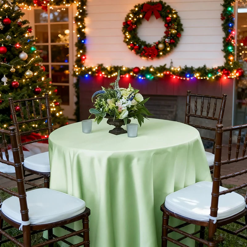 A round satin tablecloth in light green set up outdoors with wooden chairs and holiday decorations in the background