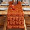 An orange textured table runner with carved pumpkin designs is displayed on a wooden table set for a holiday meal.